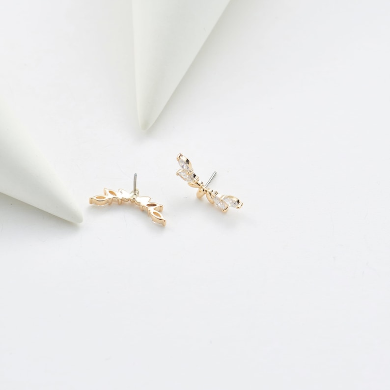 Puede incluir: Un par de trepadores de oreja dorados con un dise&ntilde;o de hojas. Cada pendiente presenta una serie de peque&ntilde;os cristales transparentes con corte marquesa. Los pendientes est&aacute;n sobre un fondo blanco, con dos formas geom&eacute;tricas blancas en el fondo.