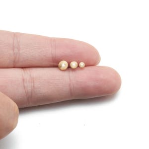 May include: Three small, gold-colored, round beads with a textured, radial pattern are displayed on a person's finger. The beads vary in size, with the largest measuring approximately 0.25 inches in diameter.