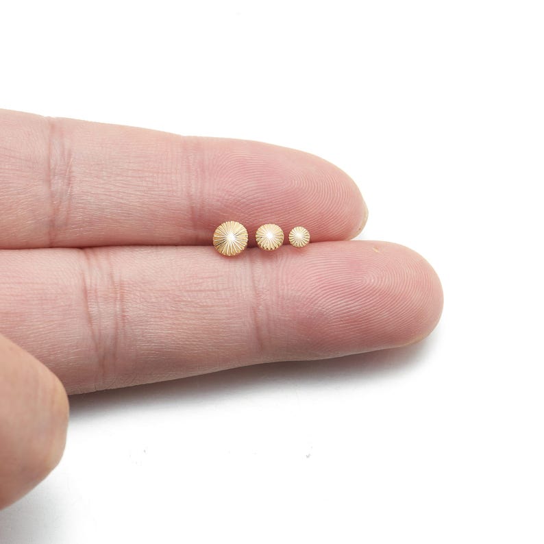 May include: Three small, gold-colored, round beads with a textured, radial pattern are displayed on a person's finger. The beads vary in size, with the largest measuring approximately 0.25 inches in diameter.
