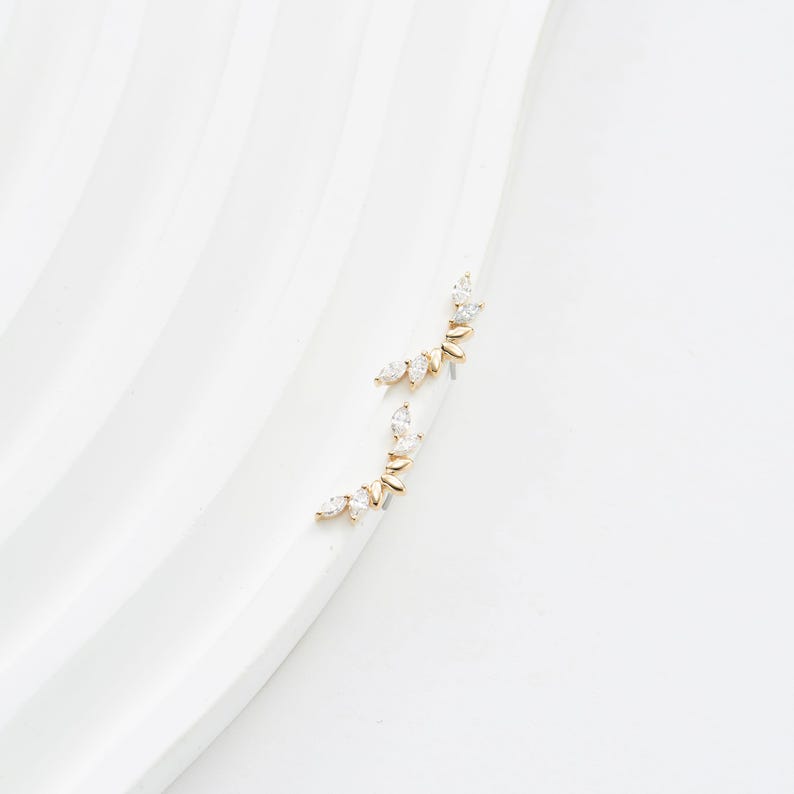 Puede incluir: Un par de trepadores de oreja dorados con un dise&ntilde;o delicado. Cada trepador presenta una serie de piedras preciosas claras con corte marquesa engastadas a lo largo de una barra curva. Los pendientes se muestran sobre una superficie blanca y curva.