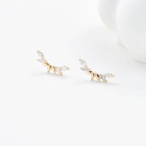 Puede incluir: Un par de pendientes trepadores dorados con un dise&ntilde;o en forma de hoja. Cada pendiente presenta m&uacute;ltiples gemas claras en forma de l&aacute;grima. Los pendientes est&aacute;n colocados sobre un fondo blanco, mostrando sus delicados detalles.