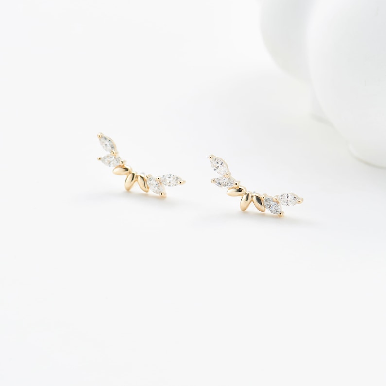 Puede incluir: Un par de pendientes trepadores dorados con un dise&ntilde;o en forma de hoja. Cada pendiente presenta m&uacute;ltiples gemas claras en forma de l&aacute;grima. Los pendientes est&aacute;n colocados sobre un fondo blanco, mostrando sus delicados detalles.