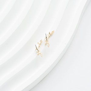 Puede incluir: Dos pendientes dorados con forma de hoja. Cada pendiente tiene una serie de hojas peque&ntilde;as y delicadas dispuestas a lo largo de un tallo curvo. Los pendientes se muestran sobre una superficie blanca texturizada.