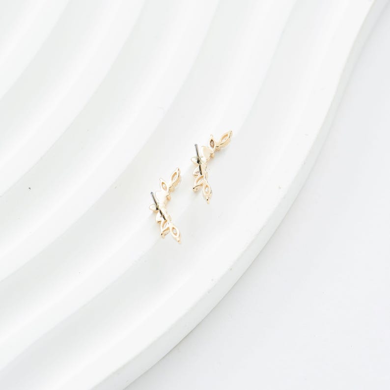 Puede incluir: Dos pendientes dorados con forma de hoja. Cada pendiente tiene una serie de hojas peque&ntilde;as y delicadas dispuestas a lo largo de un tallo curvo. Los pendientes se muestran sobre una superficie blanca texturizada.