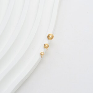 May include: Three gold-colored earrings with a sunburst design. The earrings are arranged in a vertical line, with the largest at the top and the smallest at the bottom. The earrings are on a white, textured surface.