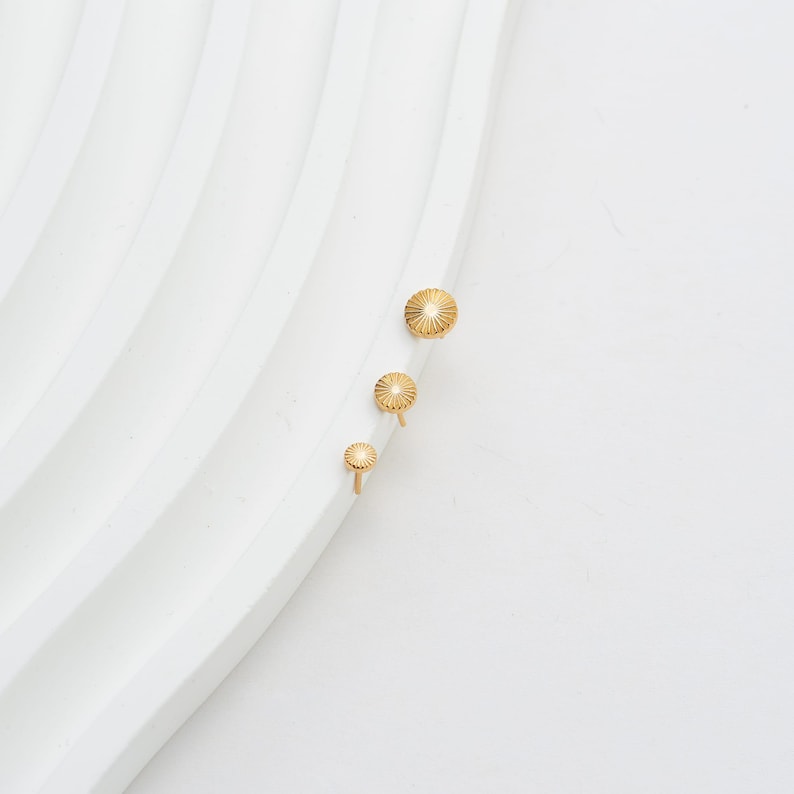 May include: Three gold-colored earrings with a sunburst design. The earrings are arranged in a vertical line, with the largest at the top and the smallest at the bottom. The earrings are on a white, textured surface.