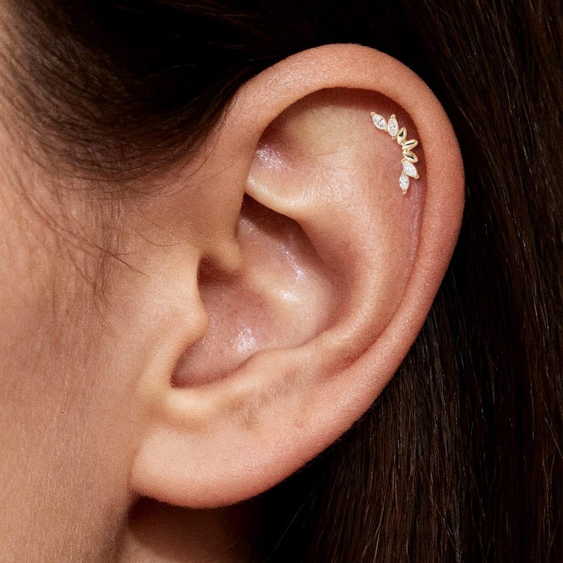 Puede incluir: Primer plano de una oreja con un piercing h&eacute;lix dorado. El piercing presenta un dise&ntilde;o curvo con varias piedras preciosas claras en forma de l&aacute;grima. La oreja es de tono claro y el fondo es oscuro.