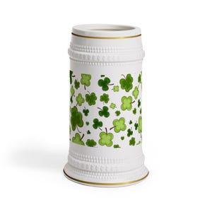 Op de afbeelding: Witte keramische bierpul met gouden rand en voet. De pul is versierd met een patroon van groene klavers in verschillende tinten. Het ontwerp herhaalt zich rond de body, wat een feestelijke uitstraling creëert.