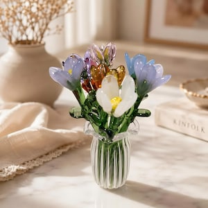 May include: A glass flower arrangement in a small, clear vase. The bouquet features colourful glass flowers in shades of blue, purple, orange, and white with green stems. The vase has a ruffled top and vertical lines.