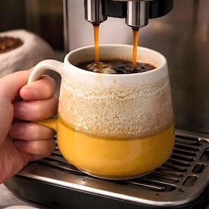 May include: A two-toned ceramic mug being filled with coffee from a coffee machine. The mug is yellow on the bottom and white with a textured, speckled design on the top. The coffee stream is dark brown.