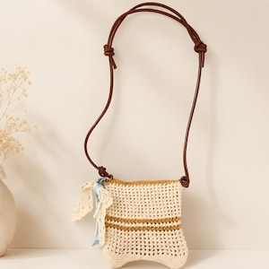 May include: A small, rectangular crochet crossbody bag in cream with two horizontal brown stripes. It features a long, thin brown cord strap with knotted ends and a decorative side accent consisting of a light blue ribbon and cream lace.