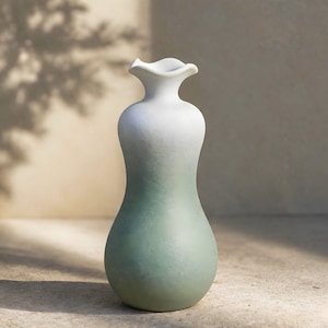 May include: A tall, elegant vase with a unique shape and a two-tone color scheme. The top of the vase is white, transitioning to a soft green at the base. The rim has a ruffled design. The vase is made of ceramic and is suitable for home decor.