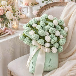 May include: A knitted bouquet of green and white pom-poms in a light green vase tied with a cream ribbon. The bouquet sits on a vintage chair. The background includes a table with flowers and a cake.