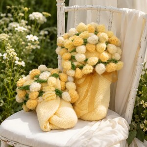 May include: Two handmade yellow and white pom-pom bouquets in knitted vases, tied with yellow ribbons, sit on a white chair. The bouquets have green accents and are set against a backdrop of greenery and white flowers.