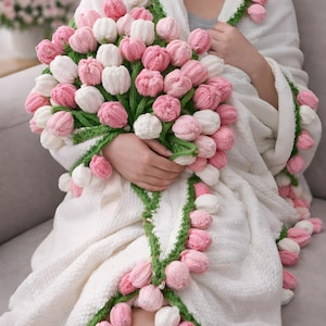 May include: A white blanket and robe adorned with crocheted pink and white tulips. The blanket is draped over a person, and a large bouquet of tulips is held in their hands. Green stems and leaves accent the floral design.