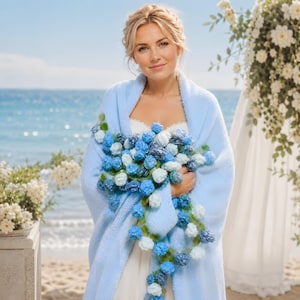 May include: A woman wearing a light blue shawl and holding a bouquet of blue and white flowers. The flowers are a mix of textures and shades, with green accents. The background features a beach and ocean scene.