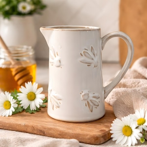 May include: Cream ceramic jug with embossed daisy patterns. The jug has a curved handle and a pouring spout. Daisies and honey are arranged on a wooden board, suggesting a rustic, farmhouse aesthetic. The jug is a decorative item.