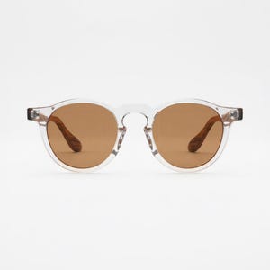 May include: Round sunglasses featuring a clear, transparent acetate frame and brown tinted lenses. The temples are constructed from a brown wood-grain material, providing a contrast to the translucent front frame and the circular lens shape.