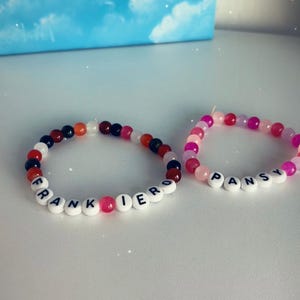 May include: Two beaded bracelets. One bracelet has the name "FRANKIERO" spelled out in white letter beads, with a mix of black, red, orange, and white beads. The other bracelet says "PANSY" with pink and purple beads.