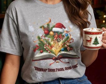 May include: A heather gray short-sleeve t-shirt featuring a graphic of an open book with a Christmas tree, ornaments, and a Santa hat emerging from the pages. The text below the graphic reads, "UNWRAP THOSE PAGES, GOOD GIRL."