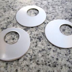 May include: Three silver-coloured, circular pendants with a central hole. The pendants are smooth and reflective, lying on a speckled grey surface. The design is simple and modern, suitable for jewellery making or craft projects.