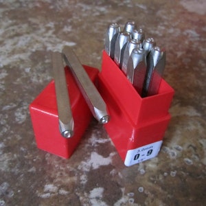 May include: A set of 10 metal letter stamps in a red plastic case. The stamps are numbered 0-9 and are 3 mm in size.