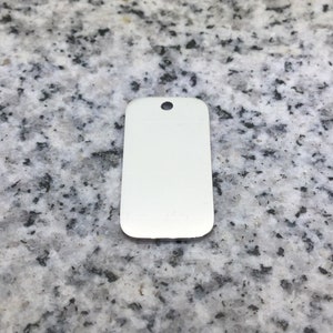 May include: A plain, white metal tag in a rectangular shape with rounded edges and a small hole at the top. It is set against a speckled grey and white surface.