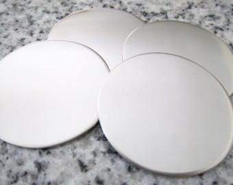 May include: Four silver-colored, round metal coasters are arranged on a speckled gray surface. The coasters have a smooth, reflective surface and a slightly raised edge. They are evenly spaced and appear to be made of a solid metal.
