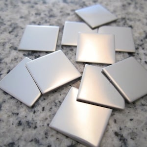 May include: A collection of silver-colored, square tiles. The tiles are arranged in a scattered pattern, with their reflective surfaces catching the light. The edges of the tiles are slightly rounded, and the overall effect is clean and modern. The tiles are likely for crafts or home decor.