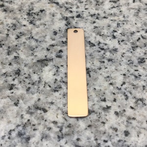 Peut inclure: Une longue pièce rectangulaire en métal mince avec un trou en haut. Le métal est de couleur or clair.