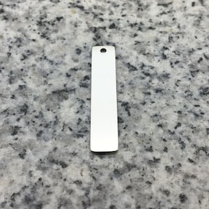 Peut inclure: Un pendentif rectangulaire argenté avec un fond arrondi et un trou en haut. Le pendentif est lisse et réfléchissant, idéal pour la gravure ou la personnalisation. Le pendentif mesure environ 3,8 cm de long.