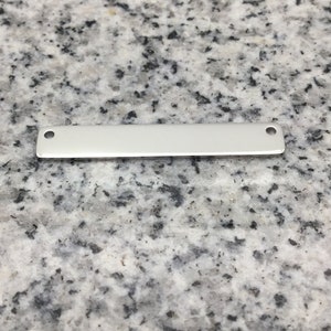 Peut inclure: Une petite étiquette rectangulaire en métal argenté avec des coins arrondis et deux petits trous, un à chaque extrémité. L'étiquette est vierge et repose sur une surface grise et blanche mouchetée.