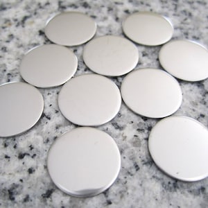May include: Ten silver-colored metal discs, each with a smooth, shiny surface. The discs are arranged in a loose circle on a speckled gray surface.