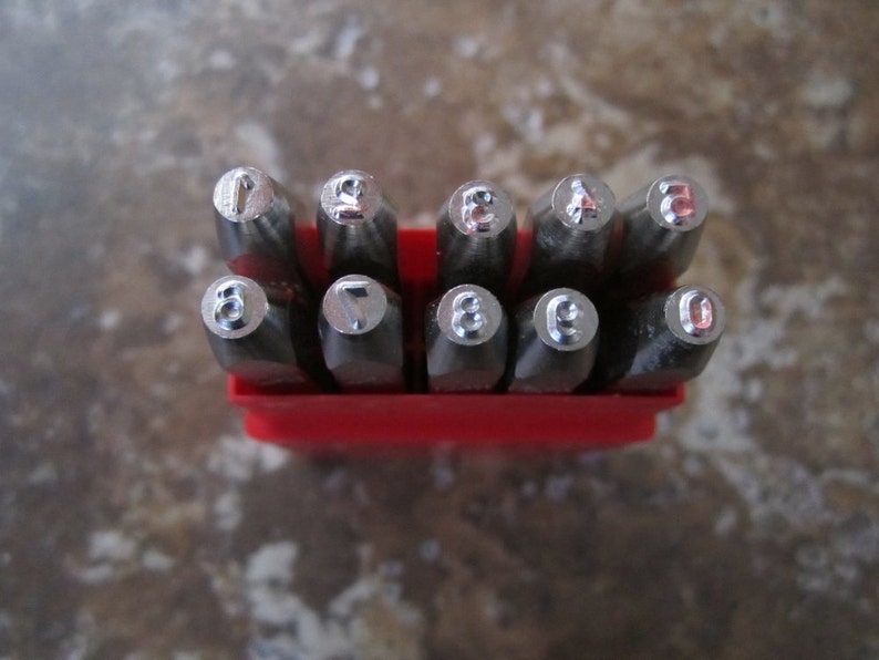 Puede incluir: Un juego de diez punzones de n&uacute;meros de metal plateado en una caja de pl&aacute;stico roja. Los punzones est&aacute;n dispuestos en orden del 1 al 0.