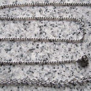 May include: A silver-colored ball chain necklace with a lobster clasp and an extender chain. The necklace is displayed on a speckled gray surface. The chain is made of small, round beads.