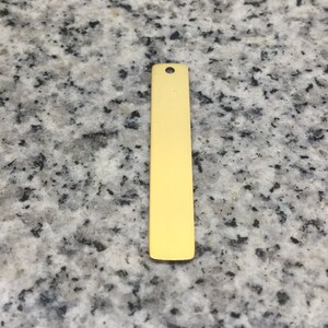 Peut inclure: Une barre en métal de couleur dorée avec un trou en haut. La barre est lisse et de forme rectangulaire.