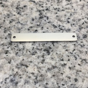 Peut inclure: Une petite étiquette rectangulaire en métal argenté avec deux trous, un à chaque extrémité. L'étiquette est vierge et repose sur une surface mouchetée grise et blanche. Idéale pour la création de bijoux ou l'artisanat.