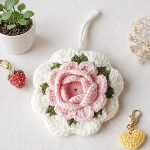 Può includere: Un sottobicchiere fatto a mano all'uncinetto con un motivo a rosa rosa, circondato da dettagli bianchi e verdi. Sono visibili anche un piccolo portachiavi a fragola e un portachiavi a cuore giallo. Gli oggetti sono su una superficie bianca.