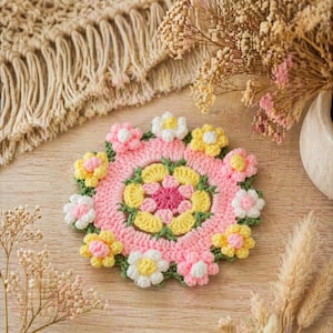 Puede incluir: Un posavasos de ganchillo hecho a mano con un diseño floral. El posavasos presenta un centro rosa con flores amarillas, blancas y rosas alrededor del borde. Hojas verdes acentúan las flores. El posavasos está sobre una superficie de madera.