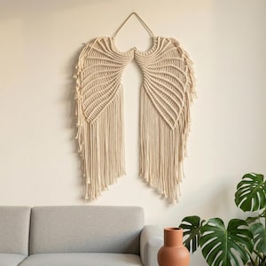 May include: A beige macrame wall hanging in the shape of angel wings. The wings are made of woven cotton rope and feature a detailed pattern of lines and fringe. The wall decor is suspended from a gold-colored metal ring.