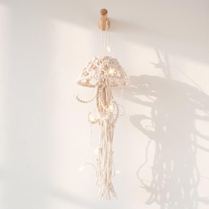 May include: A cream-colored macrame jellyfish wall hanging suspended from a wooden hook. The piece features a woven dome top with long, trailing knotted cords and is illuminated by a thin string of small, glowing LED star-shaped lights.