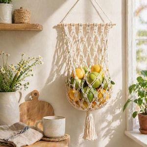 May include: A cream-colored macrame fruit basket filled with yellow and green citrus fruits. The basket hangs from a wooden dowel and has a decorative tassel. The kitchen scene includes a wooden cutting board, a mug, and a vase of flowers.
