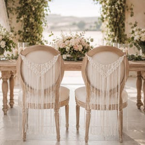 Pode incluir: Duas cadeiras de madeira com almofadas e encostos creme, adornadas com macramé. As cadeiras estão posicionadas em uma mesa de madeira decorada com flores e velas, criando um ambiente elegante para casamentos ou eventos.