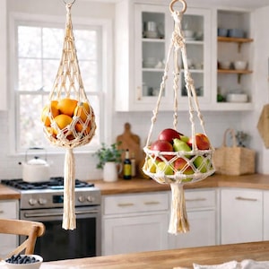 Puede incluir: Dos fruteros de macramé color crema colgando en una cocina. Uno contiene naranjas, el otro manzanas y peras. Los soportes de algodón tejido tienen una borla en la parte inferior. La cocina tiene armarios blancos y una encimera de madera.