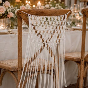 Pode incluir: Cadeira de madeira com assento e encosto tecidos, com um design de macramé em algodão creme. A cadeira tem um acabamento rústico em madeira natural, adequado para casamentos ou eventos especiais. O fundo mostra uma mesa com flores e velas.