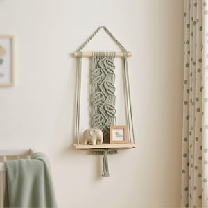 May include: A macrame wall shelf in a soft green colour, featuring a leaf design. The shelf is made of wood and holds a small elephant figurine and a framed picture. The shelf hangs from a wooden dowel and is suspended by woven cords.