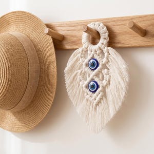 Puede incluir: Colgante de pared de macramé color crema con dos cuentas de ojo turco azules. Cuelga de un perchero de madera, junto a un sombrero de paja. Un elemento decorativo para el hogar.