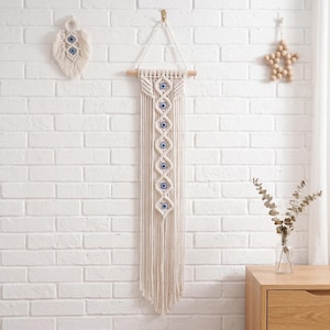Könnte beinhalten: Cremefarbener Makramee-Wandbehang mit Holzstab und Fransen. Das Design zeigt eine Reihe von blauen und weißen Evil-Eye-Perlen. Eine kleinere Makramee-Feder und Holzstern-Dekoration sind ebenfalls sichtbar.