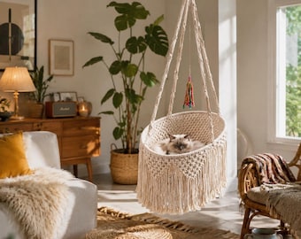 Può includere: Una sedia sospesa in macramè color crema con intricati motivi a nodo e lunghi dettagli a frangia. Un gatto soffice riposa all'interno della seduta, sospesa da spesse corde intrecciate fissate a un punto centrale superiore.