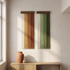 May include: Two rectangular wall hangings featuring vertical strands of fiber suspended from dark wood bars. The left piece displays a gradient of cream, yellow, and rust orange. The right piece features a gradient of cream, sage, and forest green.